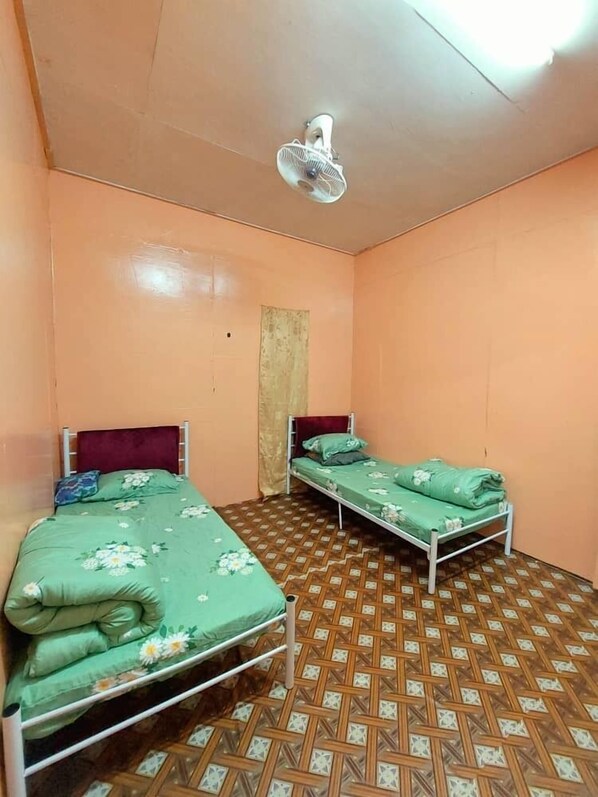 A small, single-room accommodation with two beds, a ceiling fan, and a patterned floor.