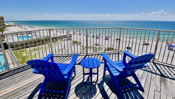 Terraza con sillas Adirondack azules y una pequeña mesa con vista a una playa con sombrillas y una piscina.