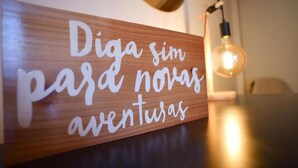 Un letrero de madera con texto cursivo blanco que dice "Diga sim para novas aventuras" en una habitación con una mesa y una bombilla colgante.