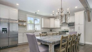 A modern kitchen with white cabinetry, a large island with a rustic table, stainless steel appliances, and a chandelier. - You Are My Sunshine (South Haven)