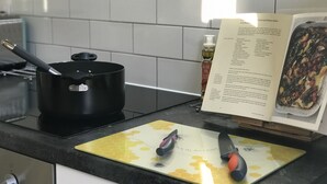 Une cuisine avec une casserole noire sur la cuisinière, une planche à découper avec des couteaux et un livre de recettes sur le comptoir.