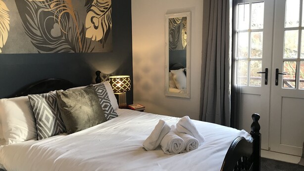 Un lit bien fait, avec du linge de lit blanc et des serviettes pliées, une tête de lit en bois foncé, une lampe fixée au mur et une fenêtre avec des rideaux.