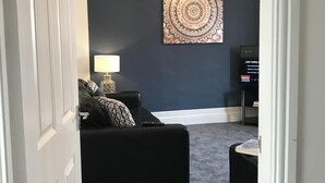 Un salon avec un mur de couleur sombre, une œuvre d’art à motifs, un canapé noir, une lampe et un téléviseur.