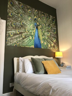 Quarto de hotel com uma grande obra de arte de pena de pavão na parede, uma cama com roupa de cama branca e um abajur.