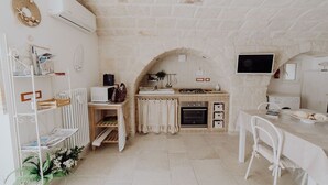 Una cocina compacta con pared de piedra, muebles blancos, una pequeña mesa de comedor y un televisor montado en la pared.