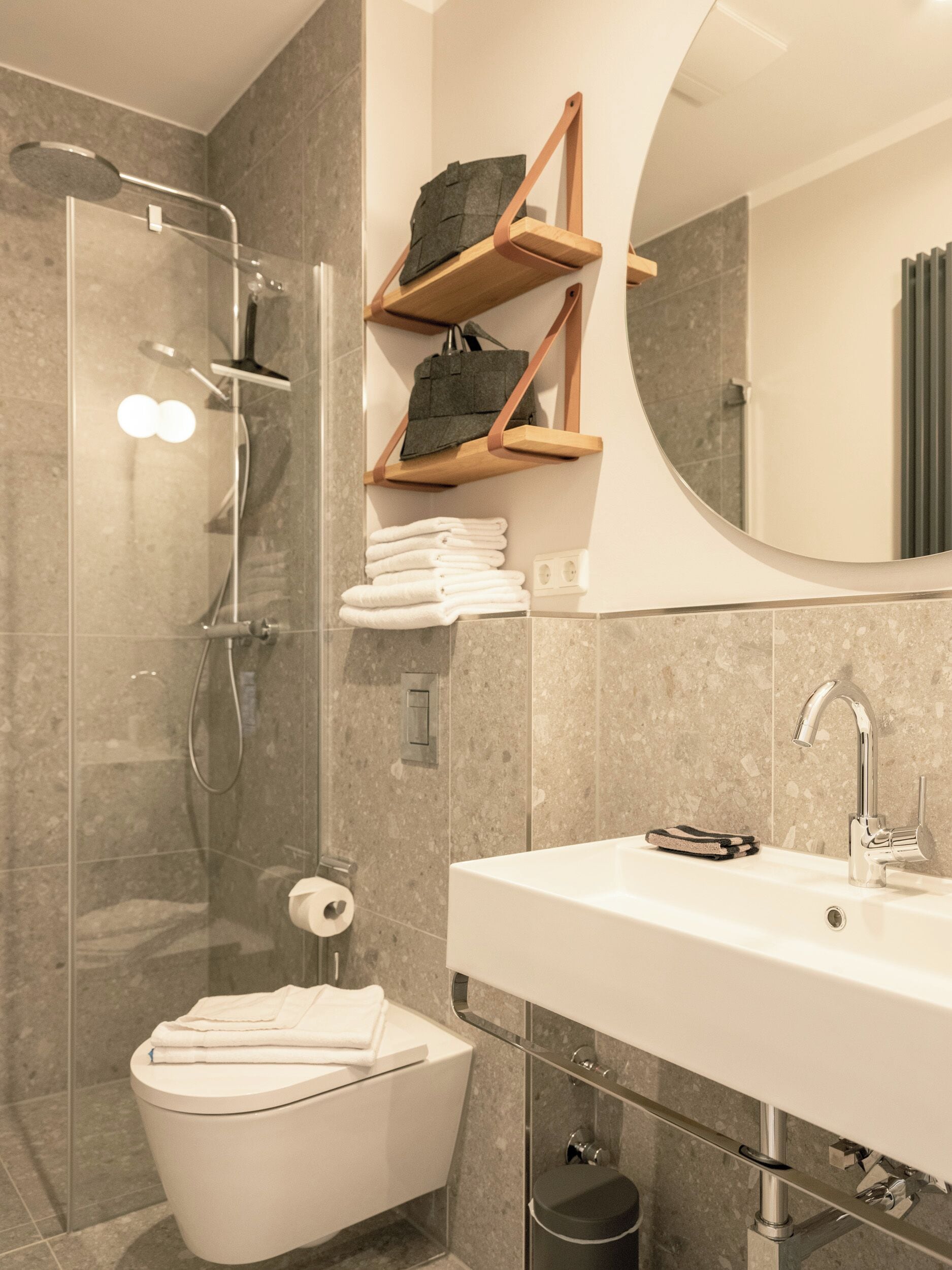 Un baño moderno con ducha de vidrio, un lavabo blanco y un inodoro. Hay toallas y un bolso en un estante de madera.