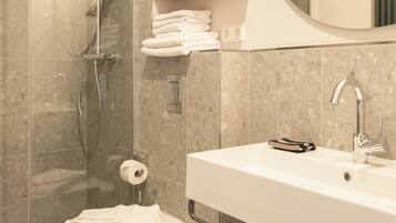 Un baño moderno con ducha de vidrio, un lavabo blanco y un inodoro. Hay toallas y un bolso en un estante de madera.
