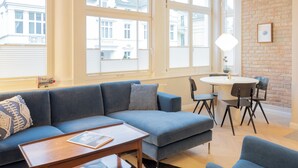 Un salon moderne avec un canapé bleu, une petite table et un coin repas avec des chaises. De grandes fenêtres offrent une vue sur la ville.