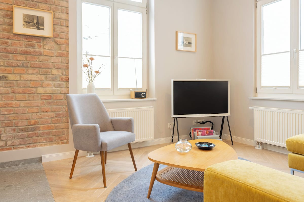 Un salón moderno con pared de ladrillo, un ventanal grande, un televisor de pantalla plana, un sillón gris, un reposapiés amarillo y una mesita con objetos decorativos.