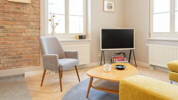 A modern living room with a brick wall, a large window, a flat-screen TV, a grey armchair, a yellow ottoman, and a small table with decorative items.