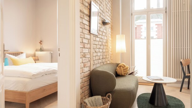 Une chambre d’hôtel moderne avec un plancher en bois, un lit recouvert de draps blancs, un fauteuil vert, une petite table ronde et un panier en osier.