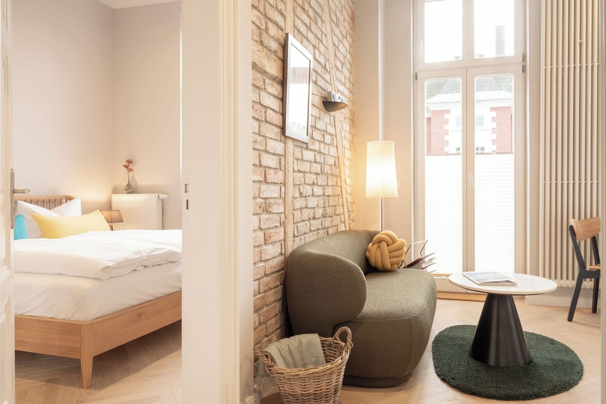Una habitación de hotel moderna con piso de madera, una cama con ropa blanca, un sillón verde, una mesita redonda y una cesta de mimbre.
