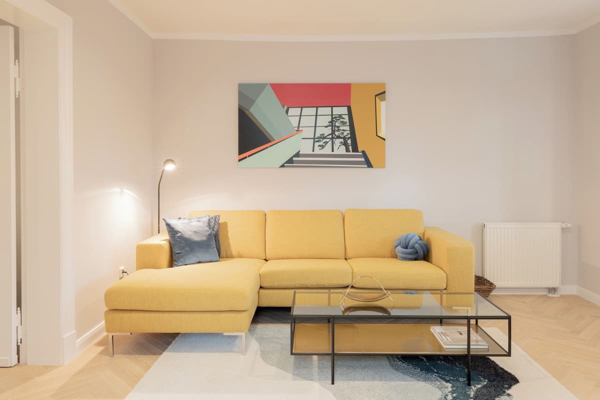 Una sala de estar moderna con un sofá amarillo, una mesa de centro de vidrio y un cuadro en la pared.