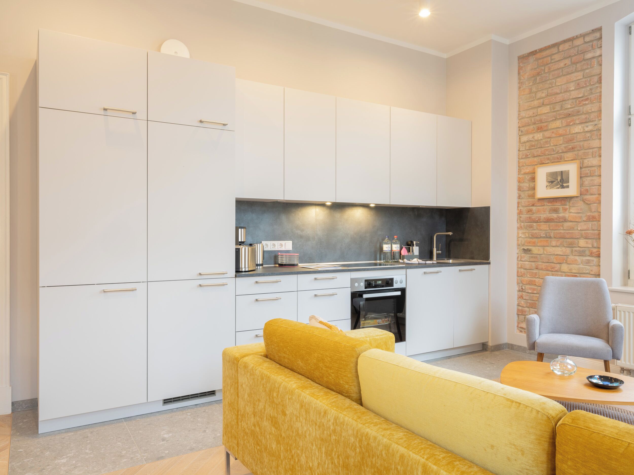 A modern kitchen with white cabinets, a black countertop, and a brick wall. There is a yellow sofa and a gray armchair in the living area.