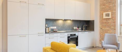 A modern kitchen with white cabinets, a black countertop, and a brick wall. There is a yellow sofa and a gray armchair in the living area.