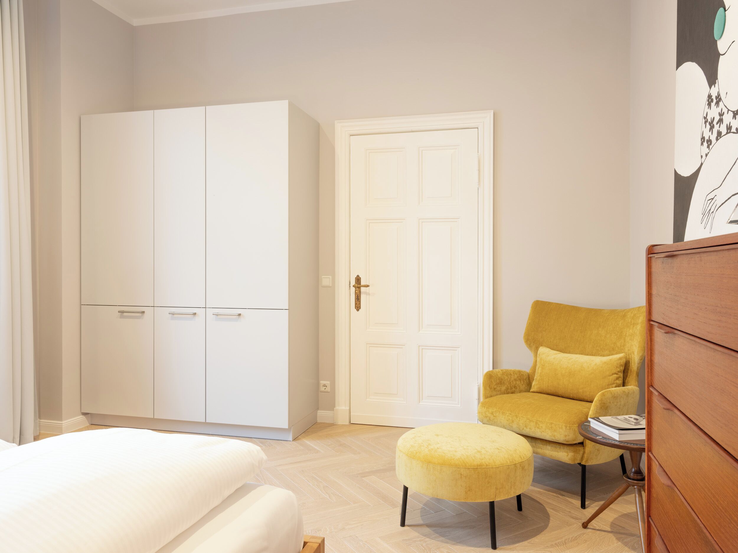 A bedroom with a bed, a wardrobe, a door, a yellow armchair, and a small table with a book.