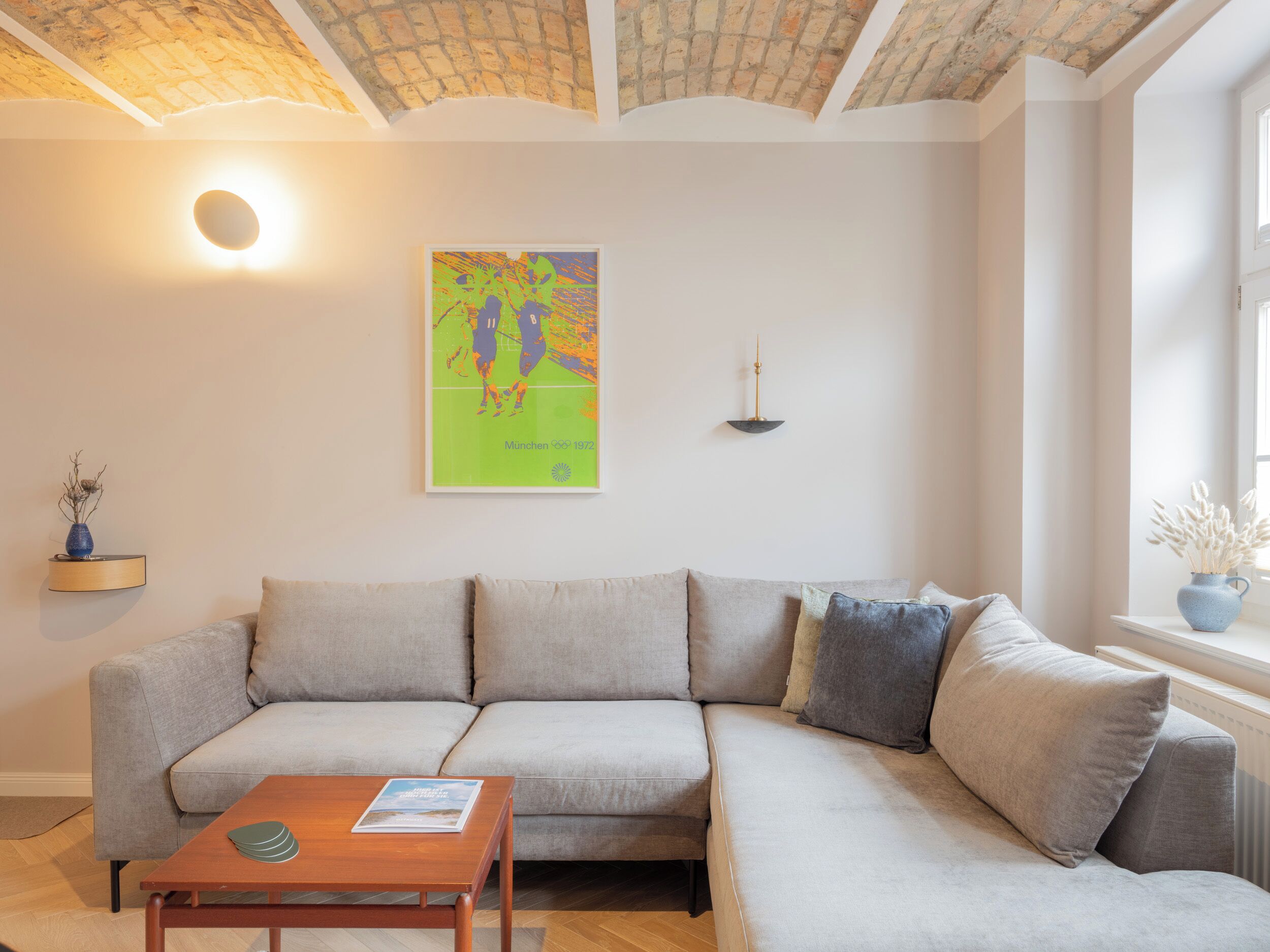 Ein modernes Wohnzimmer mit einem hellgrauen Ecksofa, einem Holztisch, einem gerahmten Kunstwerk an der Wand und einem Fenster mit Blick ins Freie.