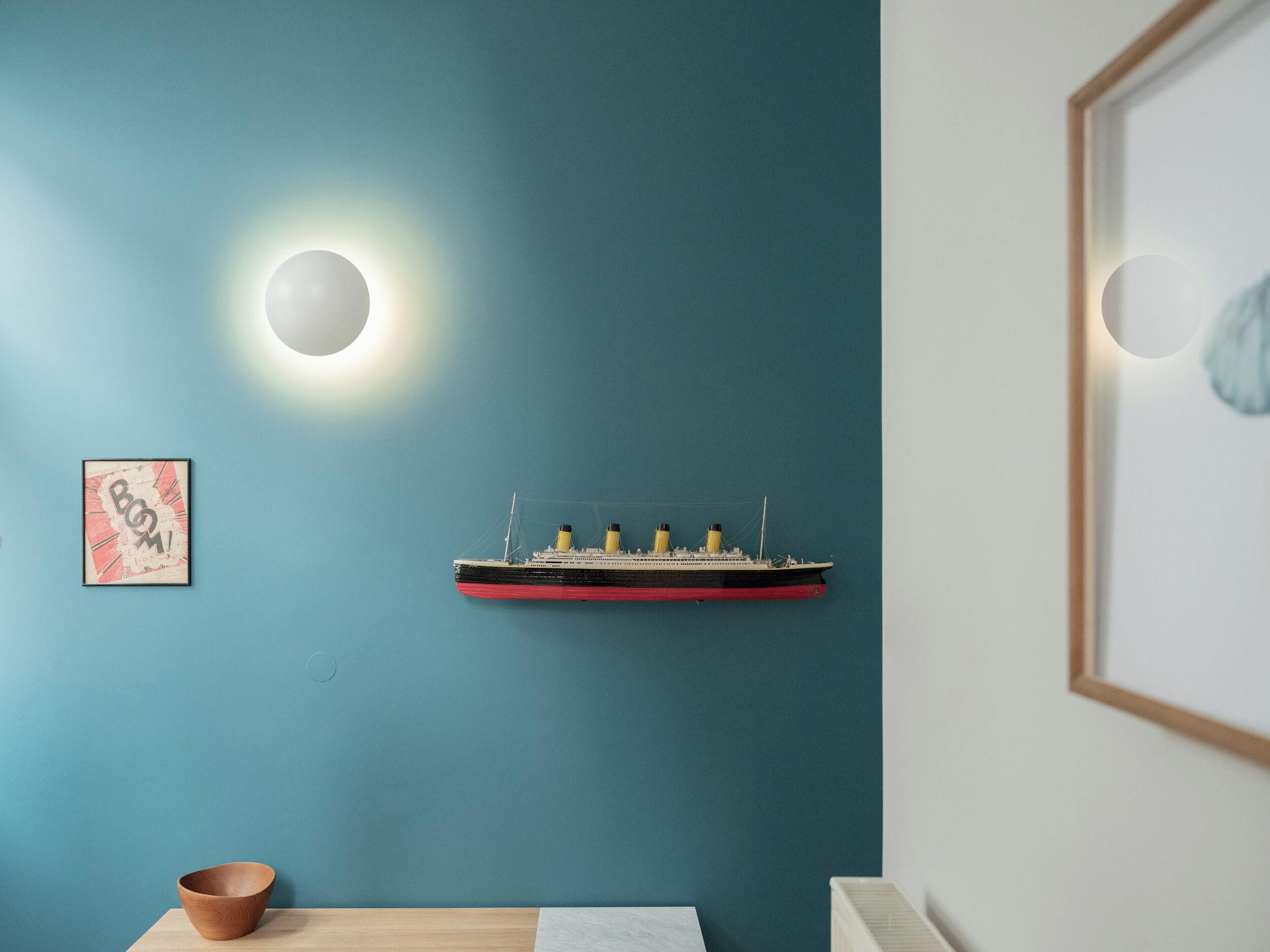 A room with a teal wall, a wooden table, a small wooden bowl, a framed picture, and a model ship.