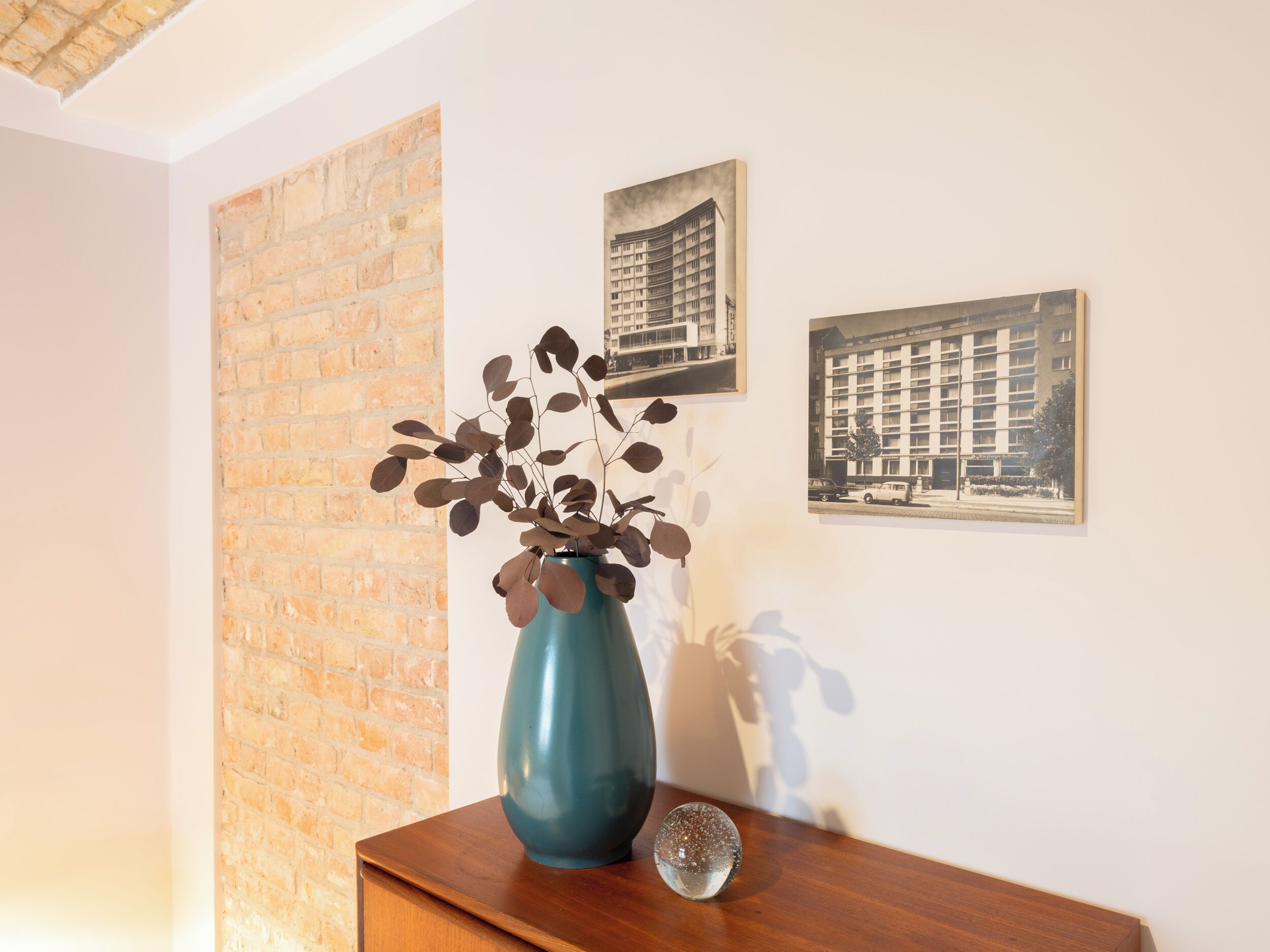 A room with a brick wall, a blue vase with dried leaves, and two framed pictures on the wall.