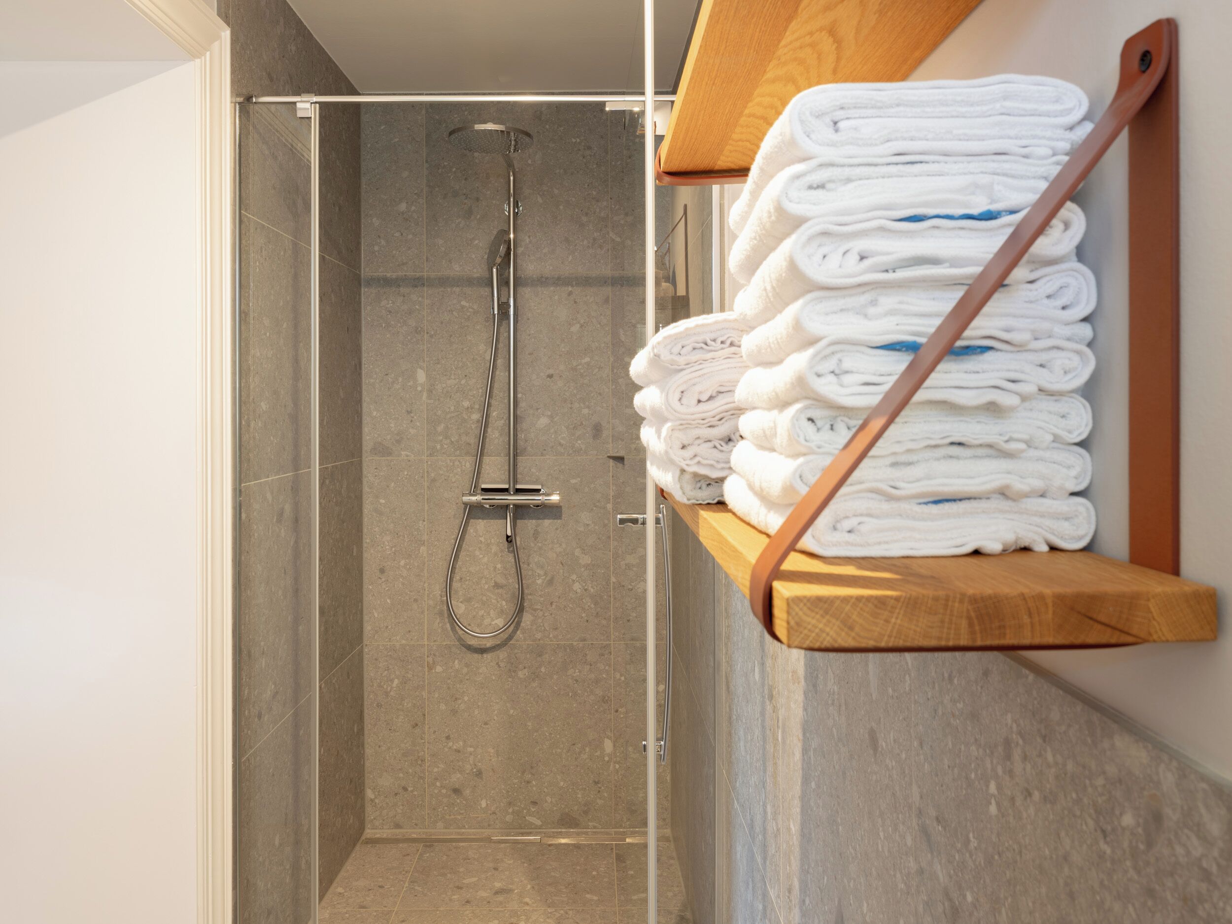 A shower area with a glass door, a metal showerhead, and a wooden shelf holding folded towels.