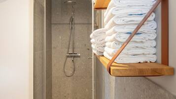 A shower area with a glass door, a metal showerhead, and a wooden shelf holding folded towels.