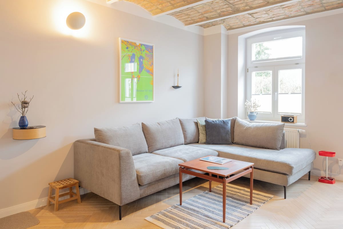 Una sala de estar moderna con un sofá gris, una pequeña mesa de madera y un gran ventanal que proporciona luz natural.