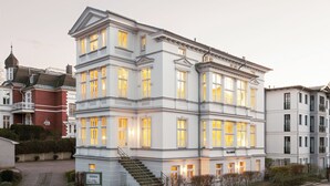 Un immeuble à plusieurs étages de style architectural classique, doté d’une entrée imposante avec un escalier et une enseigne sur laquelle on peut lire « ILLA GROTH ».