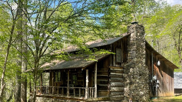 A rustic log cabin with a stone chimney, surrounded by dense trees. - Cabins In The Pines Unit 2-Covered Porch -  - Charcoal Grill - Wood Fireplace (Cherokee)