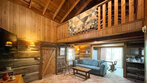A rustic cabin interior with a wooden ceiling, a large quilt hanging on the wall, a cozy seating area with a sofa and chairs, and a kitchen visible through an open door. - Cabins In The Pines Unit 2-Covered Porch -  - Charcoal Grill - Wood Fireplace (Cherokee)