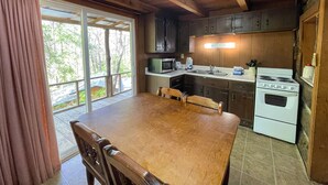 A wooden cabin kitchen with a dining table, cabinets, a stove, and a large window with curtains. - Cabins In The Pines Unit 2-Covered Porch -  - Charcoal Grill - Wood Fireplace (Cherokee)