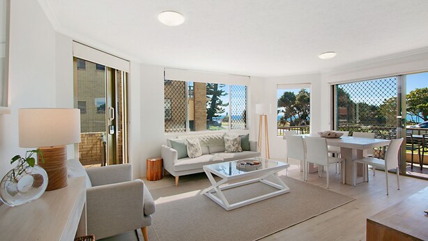 A modern living room with a sofa, coffee table, and dining area. Large windows provide views of the outdoors. - Point View Lodge 2 - Ground Floor Beachfront Bilinga - Linen Provided (Bilinga)