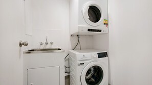 A compact laundry room with a washing machine and dryer stacked vertically, a sink with a faucet, and a door with a round knob. - Point View Lodge 2 - Ground Floor Beachfront Bilinga - Linen Provided (Bilinga)