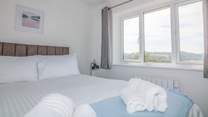 A neatly made bed with white linens and a blue accent, a bedside lamp, and a window with a view of the landscape. - Waters Reach (Kingsbridge)