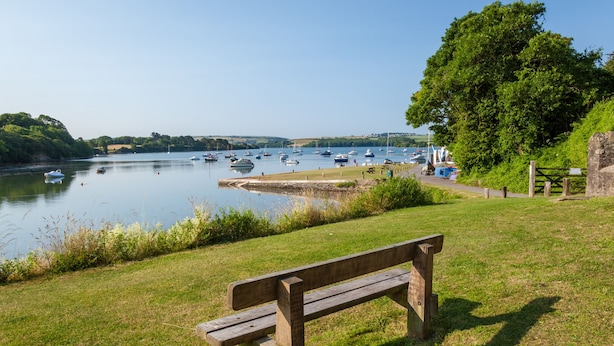 A wooden bench overlooking a calm lake with boats, a grassy area, and a stone wall. - Waters Reach (Kingsbridge)