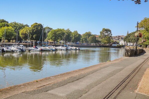 A marina with boats docked along the water's edge, a paved walkway, and trees in the background.