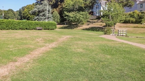A grassy field with a path leading to a building. There are trees and a bench in the background. - Waters Reach (Kingsbridge)