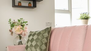 En rosa soffa med en grön- och vitrutig kudde, en vas med blommor och en svart hylla med små växter.