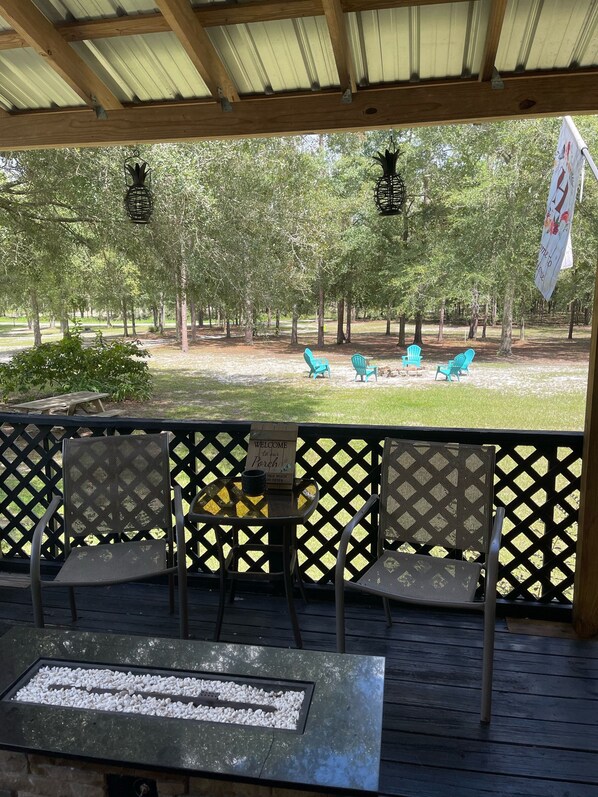A covered outdoor seating area with metal chairs and a table, overlooking a grassy park with trees and a flag. - Cave Divers Spring Escape, Sleeps 8+, Private Patio, Fire Pit, Spacious Kitchen (McAlpin)