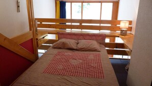 Ein Einzelbett mit karierter Bettdecke und Kissen, ein hölzernes Kopfende, ein Nachttisch mit Lampe sowie ein Fenster mit Vorhängen.