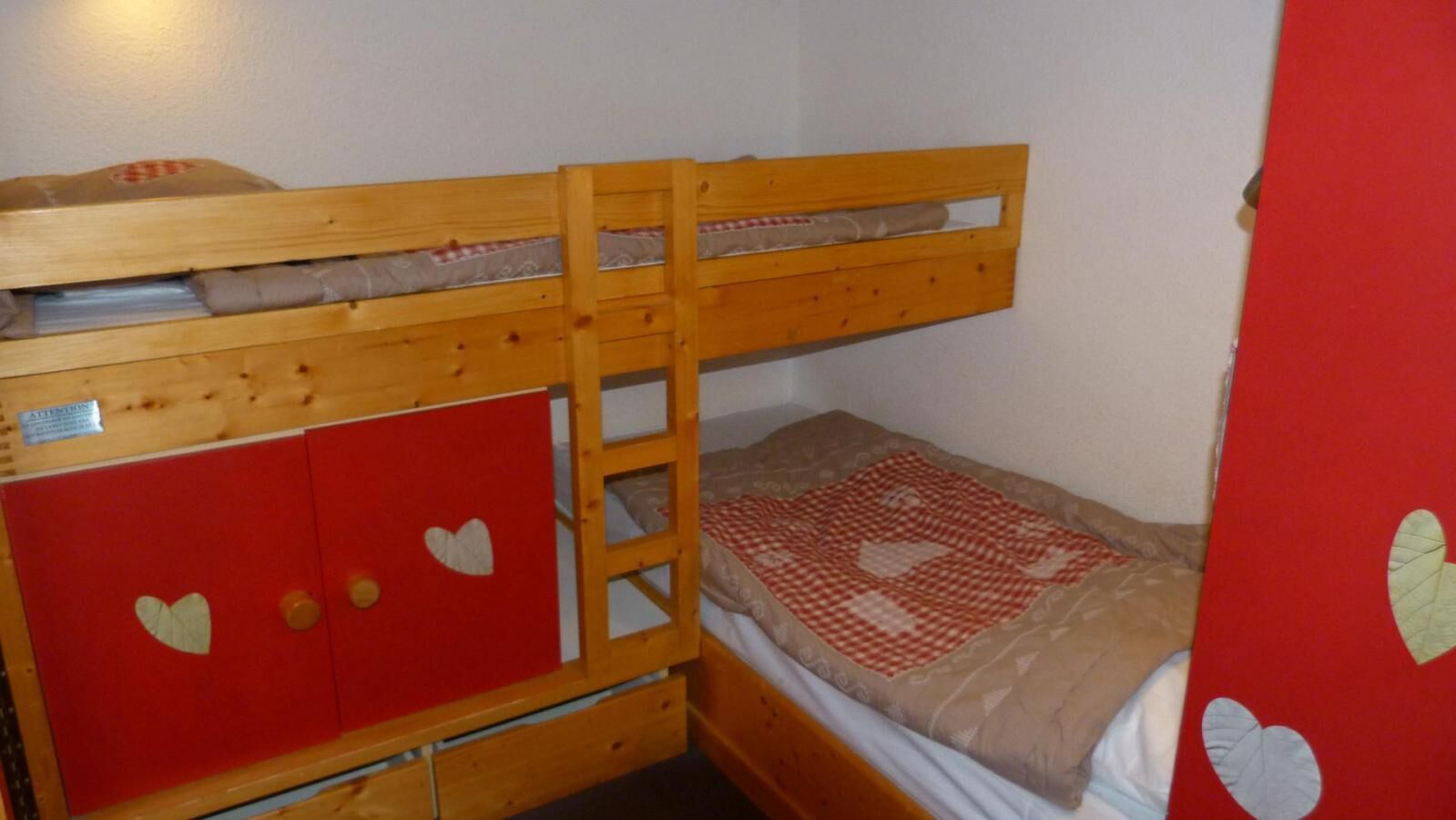 A bunk bed with a ladder, a red wardrobe with heart cutouts, and a patterned bedspread.