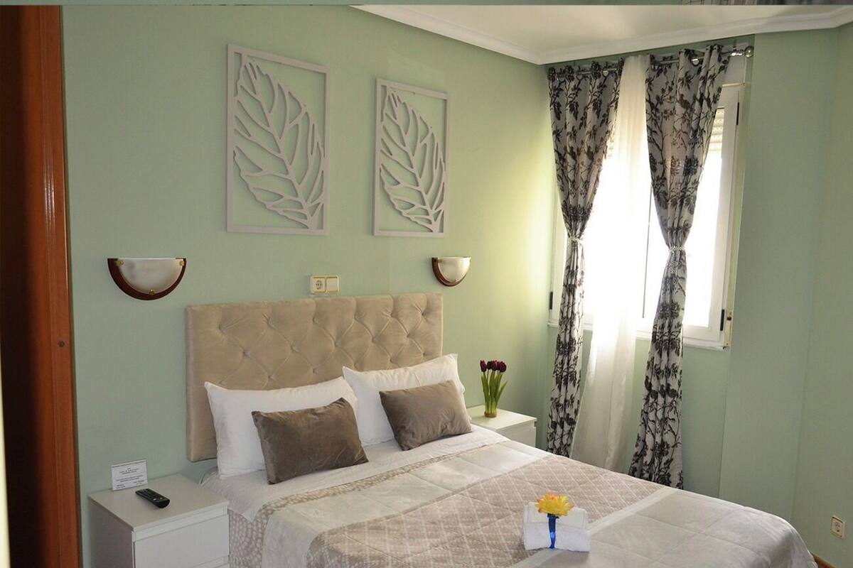 Une chambre à coucher avec un lit, une table de chevet, des œuvres d’art murales, des rideaux et un vase avec des fleurs.