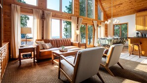 A cozy cabin interior with a wooden ceiling, large windows, a dining area, and a seating area with a sofa and chairs.