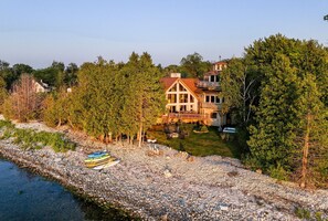 A large house with a prominent porch, surrounded by dense trees and a rocky shoreline.