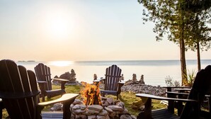A serene outdoor seating area with Adirondack chairs and a stone fire pit, overlooking a calm body of water during sunset.