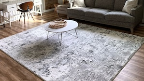 Un salon avec un canapé gris, une table basse blanche, un tapis à motifs, une table à manger en bois avec des chaises, et une grande fenêtre avec des rideaux.