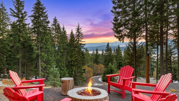 Una fogata circular rodeada de sillas Adirondack rojas en un entorno boscoso, con vistas a montañas y al atardecer.