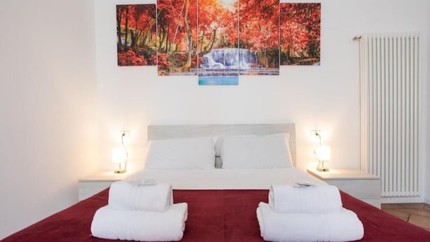 A hotel room with a large bed, two bedside tables with lamps, a red carpet, and a multi-panel artwork above the bed.