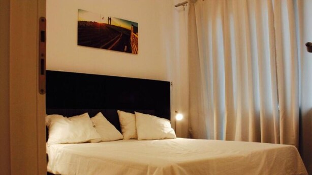 A neatly made bed with a dark headboard, a single wall-mounted picture, and a window with curtains. - Apartment Central Gardens (București)