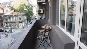 A balcony with a view of a city street, a small table with chairs, and an air conditioning unit on the wall. - Apartment Central Gardens (București)