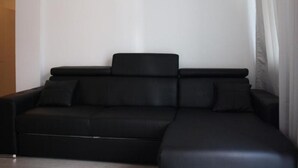 A black sectional sofa with a chaise, positioned against a plain wall with a curtain partially drawn to the side. - Apartment Central Gardens (București)